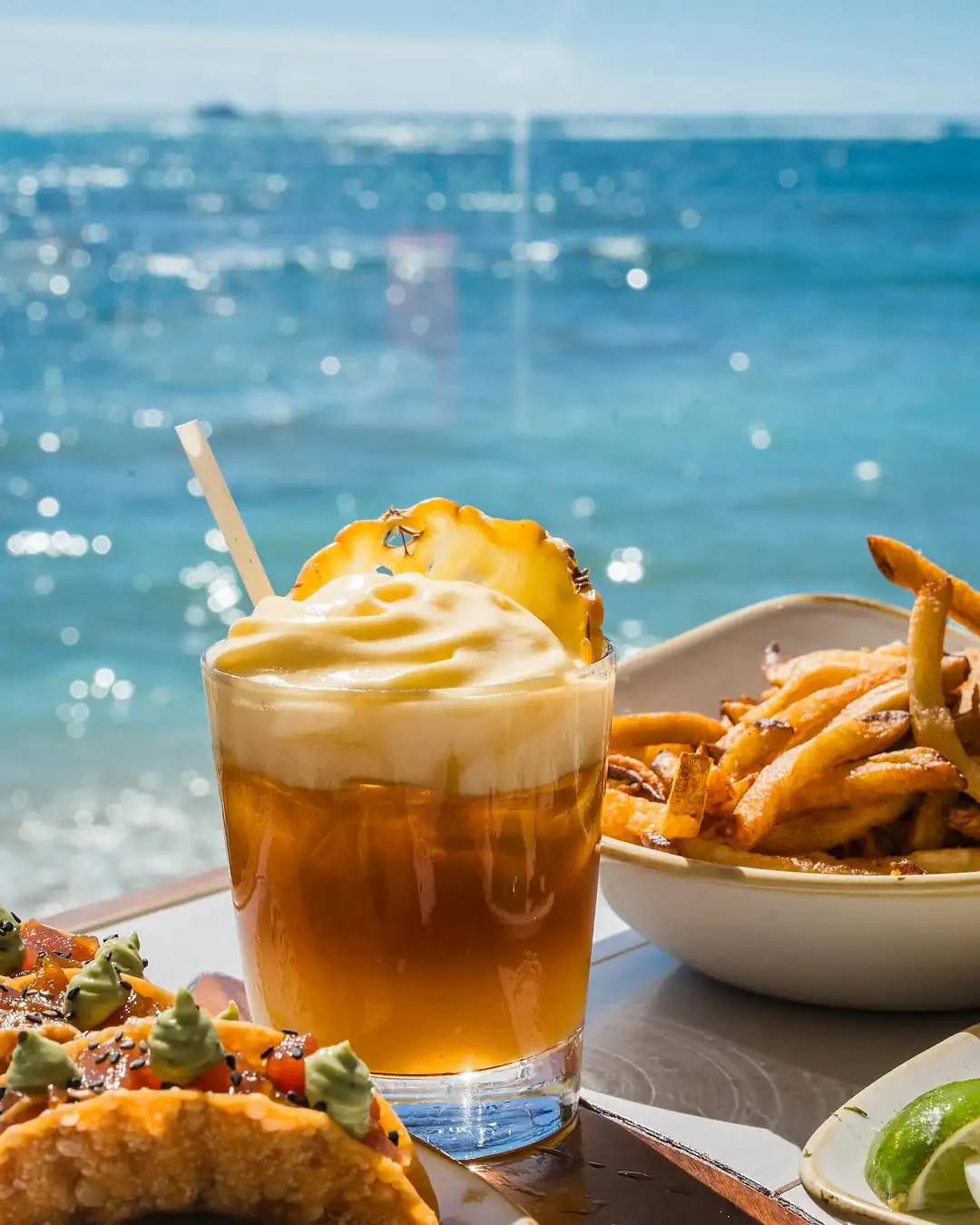 A tropical cocktail with a pineapple slice, fries, and food sit on a table by the ocean, with sunlight sparkling on the water in the background.