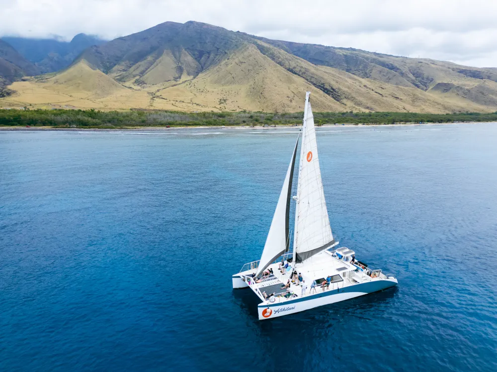 A white catamaran with raised sails floats on clear blue water near a coastline with green hills and mountains in the background.