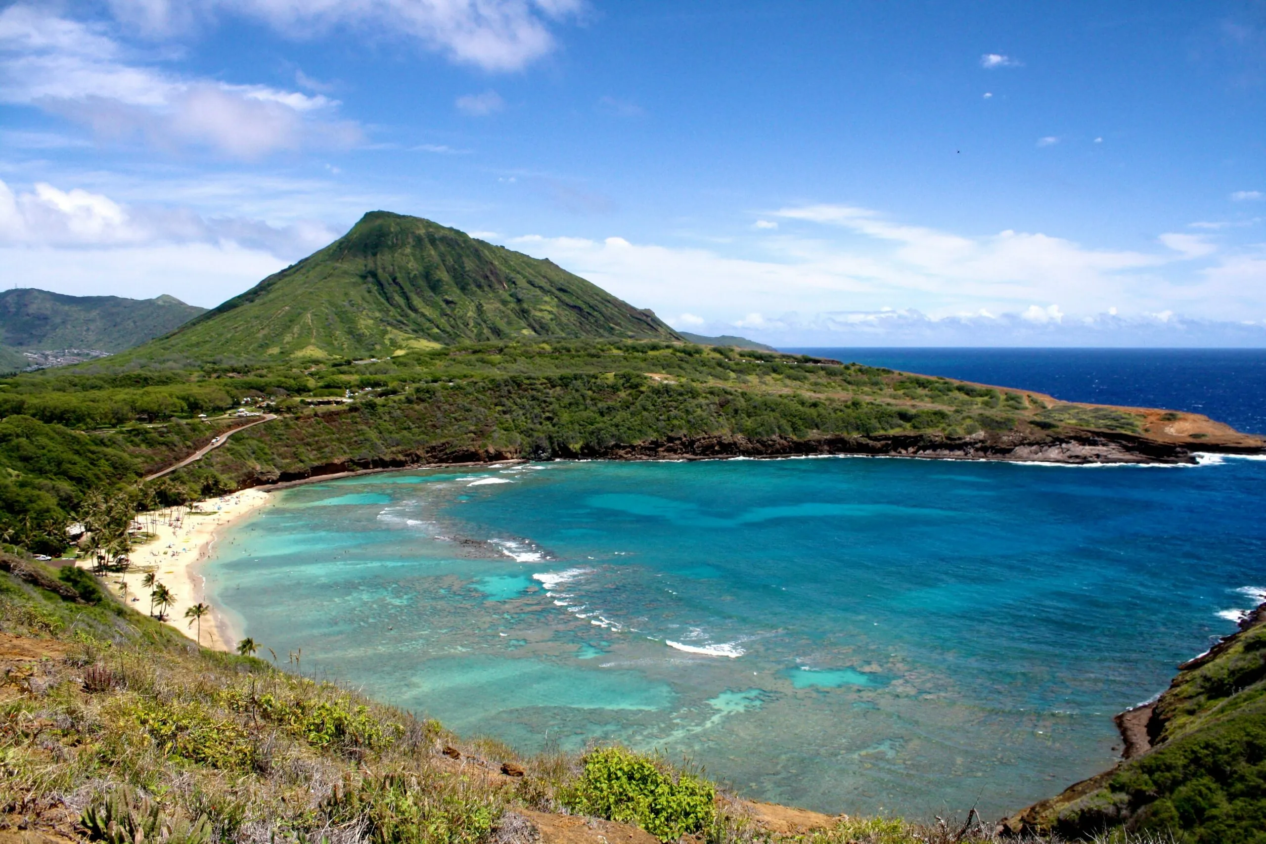 A turquoise bay with a sandy beach is surrounded by lush green hills and a prominent mountain under a blue sky with scattered clouds.
