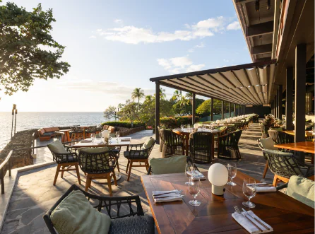 Outdoor restaurant with wooden tables and chairs overlooking the ocean, set for dining, with trees and a clear sky in the background.