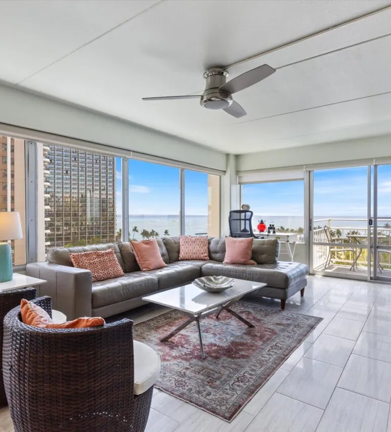Modern living room with large windows, ocean view, gray sectional sofa, patterned rug, two wicker chairs, and ceiling fan.