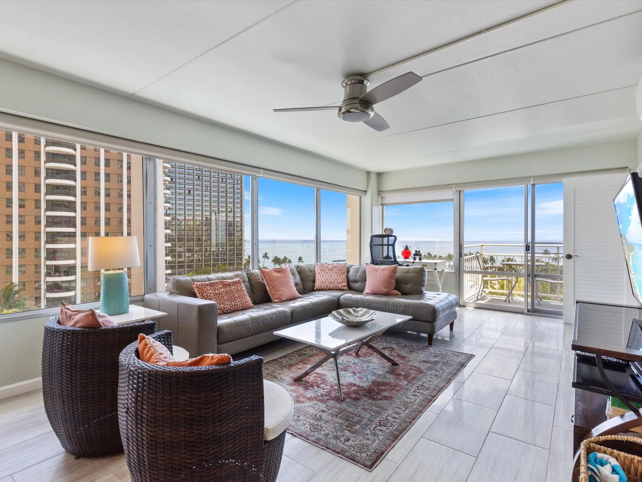 Modern living room with large windows, ocean view, gray sectional sofa, patterned rug, two wicker chairs, and ceiling fan.