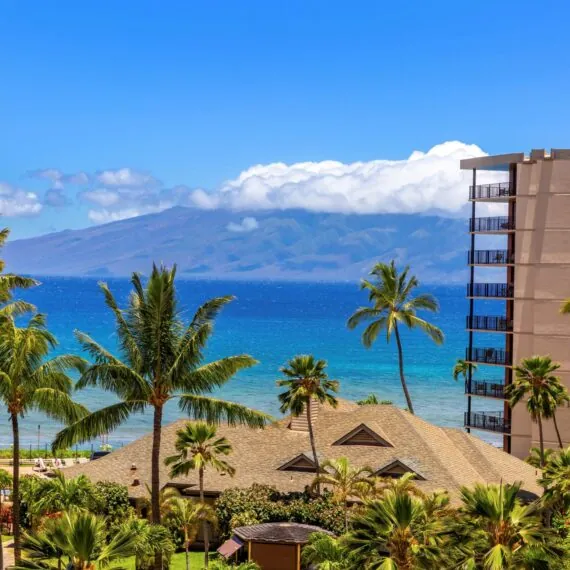 View of the ocean with palm trees, a sandy beach, and mountains in the background, next to a tall resort building under a clear blue sky.