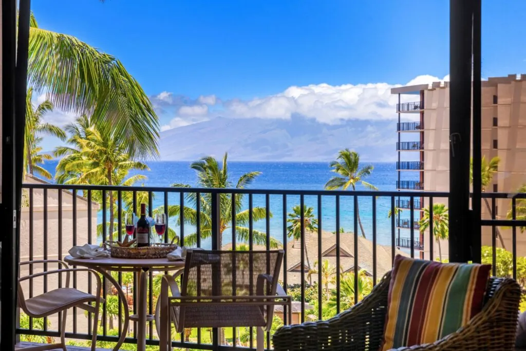 Balcony with a small table, chairs, wine, and ocean view, surrounded by palm trees and buildings under a bright blue sky with clouds.