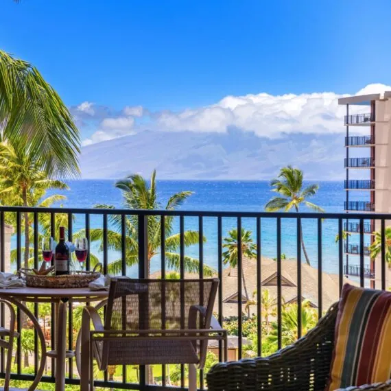 Balcony with a small table, chairs, wine, and ocean view, surrounded by palm trees and buildings under a bright blue sky with clouds.