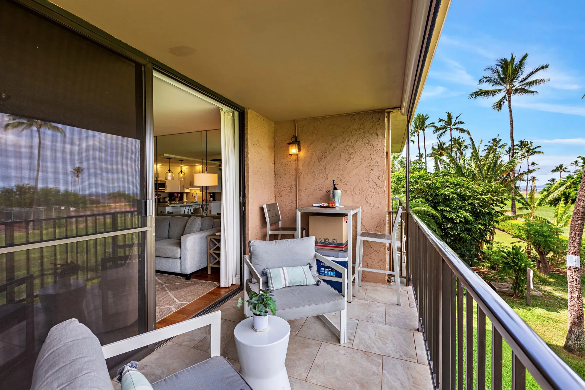 Furnished balcony with chairs and a small table, overlooking a lush green landscape with palm trees; sliding door leads to a modern living room.