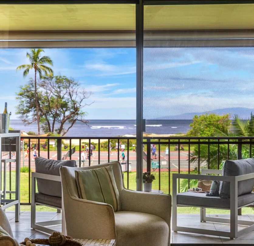 View from a living room with cozy chairs and a balcony overlooking the ocean, palm trees, and a road with people walking and jogging.