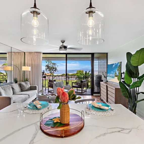 Bright, modern living room with ocean view, tropical decor, large windows, indoor plants, pendant lights, and a set dining area on a marble island.