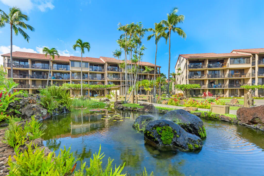 A tropical resort with palm trees, a pond with rocks and ferns, and three-story buildings with balconies under a bright blue sky.