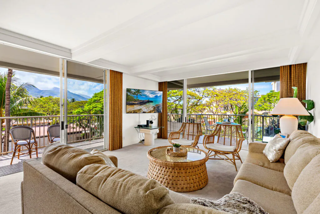 Bright living room with beige sofa, rattan furniture, large windows, balcony seating, and views of tropical trees and mountains outside.