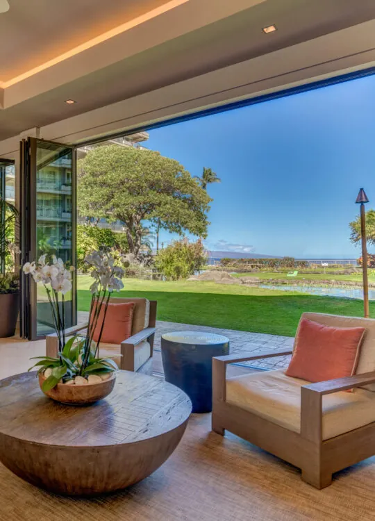 Spacious living room with modern decor opens to a lush garden and ocean view through large glass doors on a sunny day.
