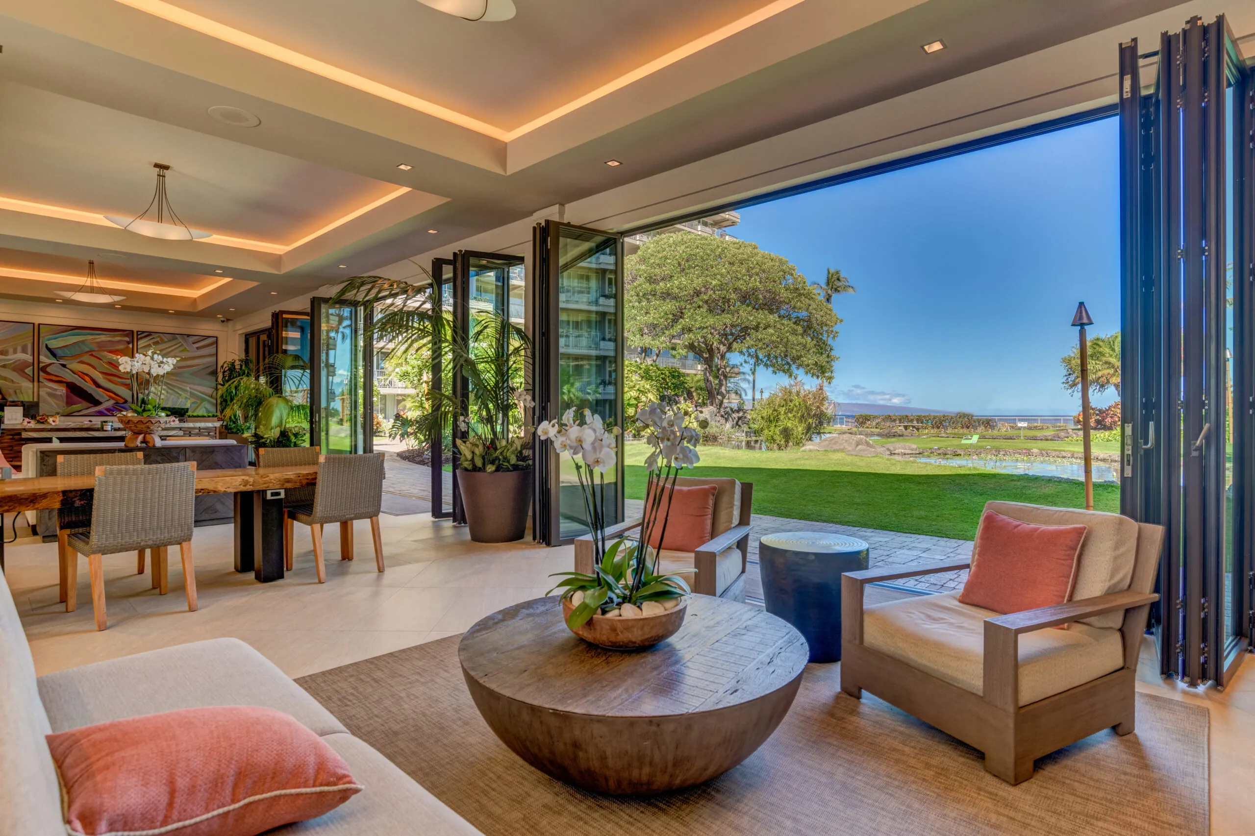 Spacious living room with modern decor opens to a lush garden and ocean view through large glass doors on a sunny day.