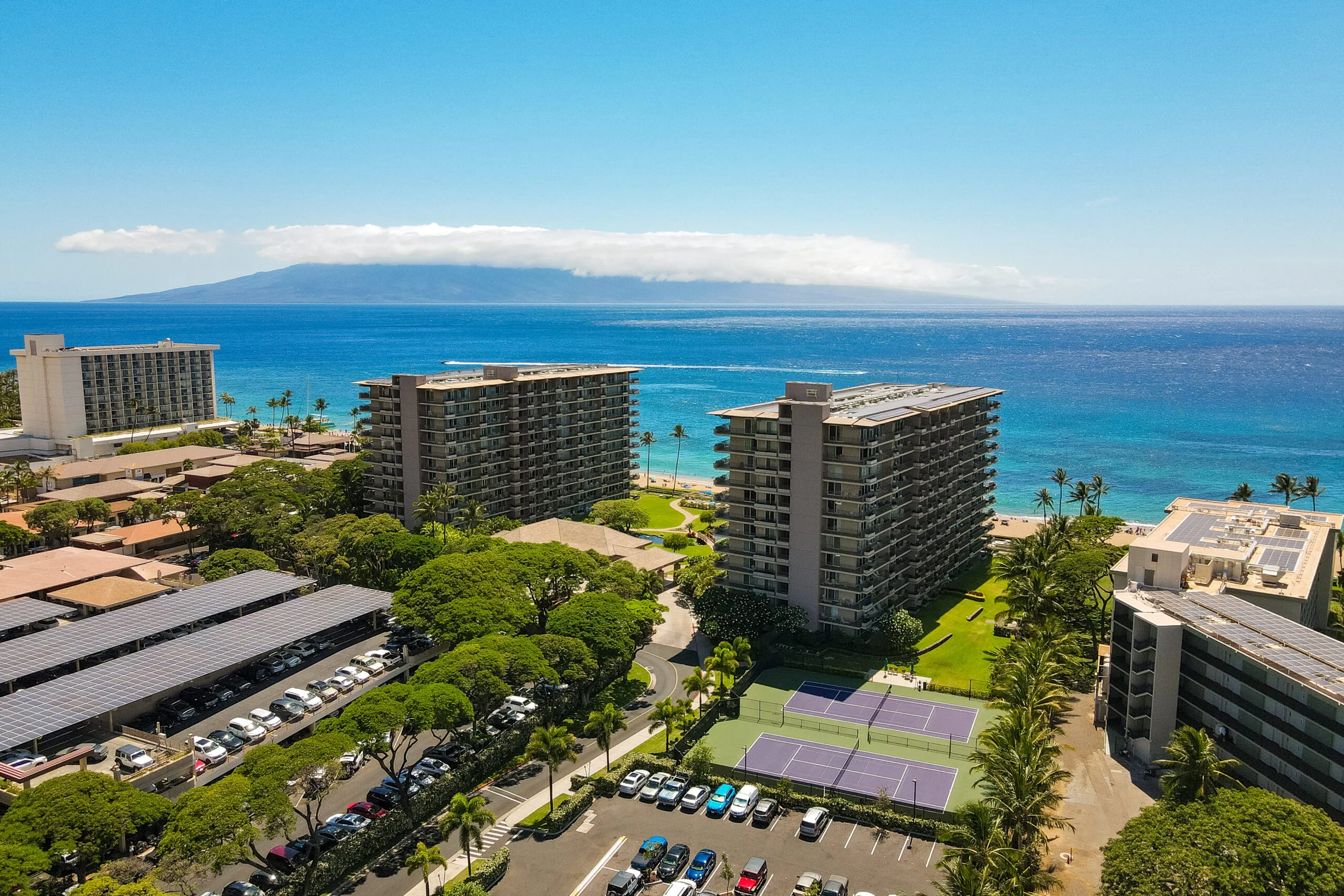 Aerial view of beachfront hotels, tennis courts, palm trees, and parked cars by the ocean, with an island and blue sky in the background.