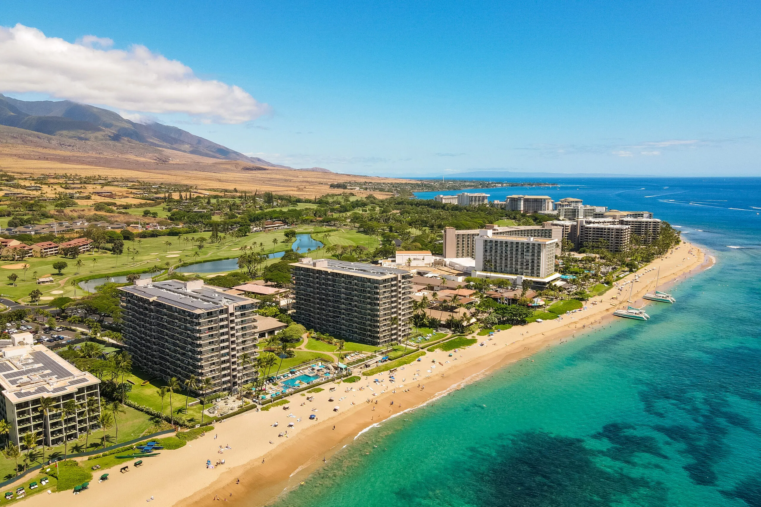 Aerial view of beachfront resorts along a sandy beach with turquoise water and mountains in the background under a clear blue sky.