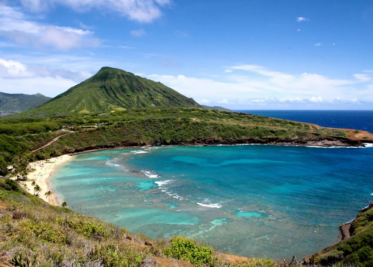 A turquoise bay with a coral reef, sandy beach, and lush green mountain under a blue sky with scattered clouds.