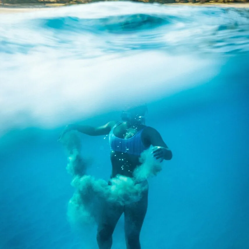 A person in a swimsuit floats underwater near the surface, with sand swirling around their legs; mountains and trees are visible above water.