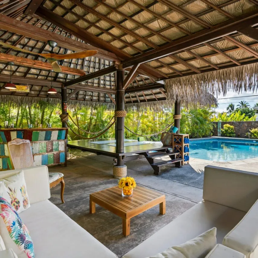 Covered outdoor lounge with sofas, a wooden table, hammock, bar area, and view of a swimming pool surrounded by tropical greenery.