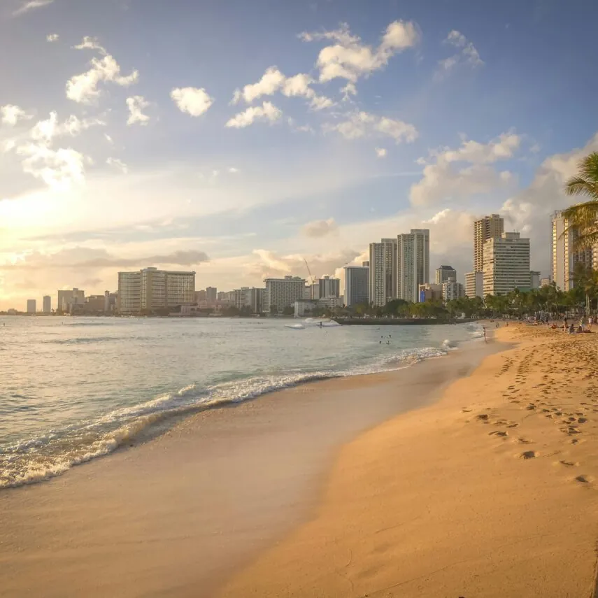 Sunny beach with gentle waves, palm trees, and footprints in the sand, with a city skyline in the background under a partly cloudy sky.