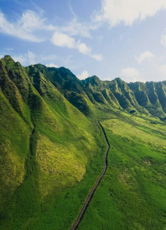 Aerial view of lush green mountains with a winding narrow path under a blue sky with scattered clouds.