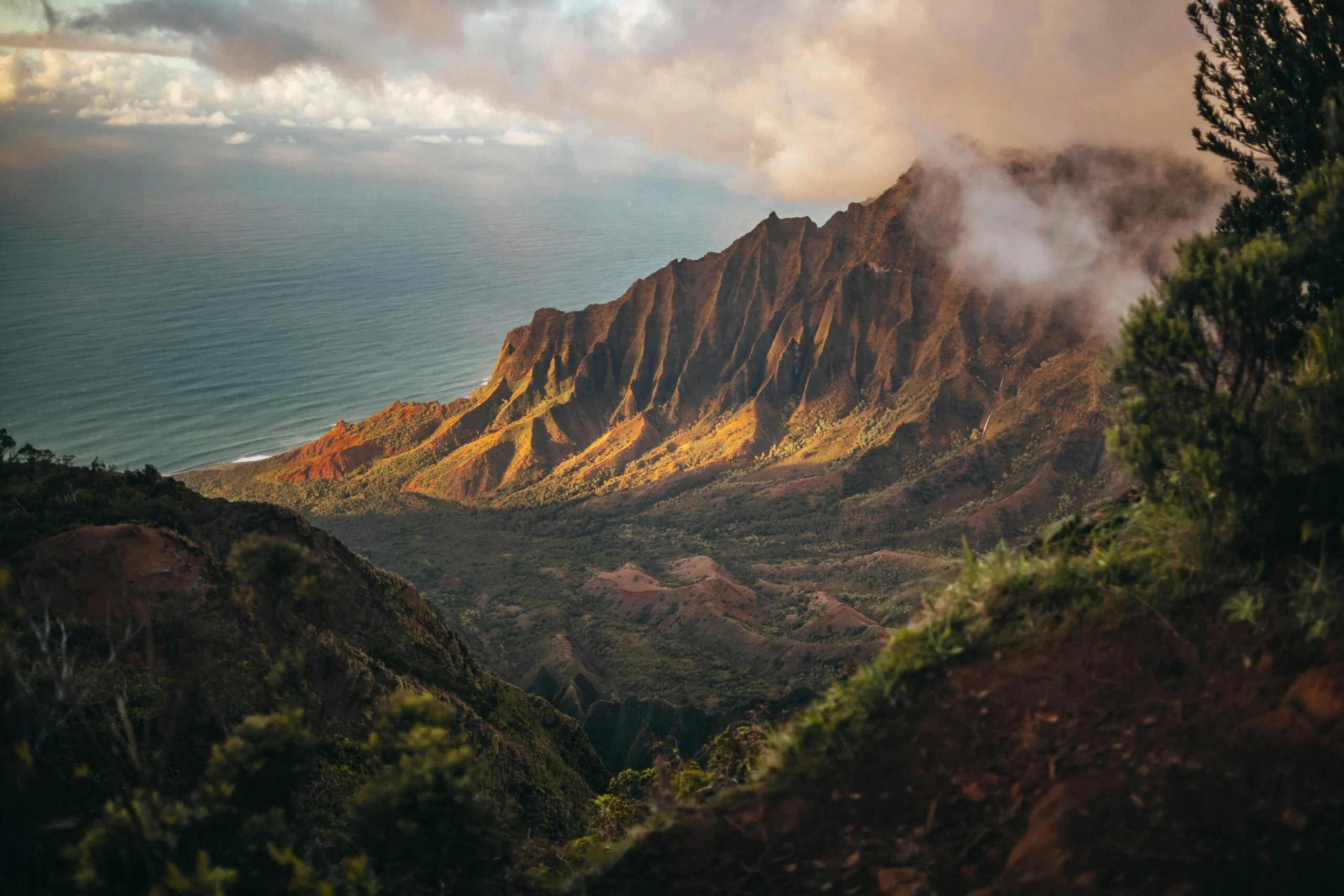 Dramatic cliffs and lush green hills overlook the ocean under a cloudy sky, with sunlight highlighting the landscape.