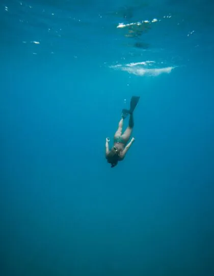 A person wearing swim fins dives underwater in clear blue ocean, surrounded by deep water and sunlight filtering from above.