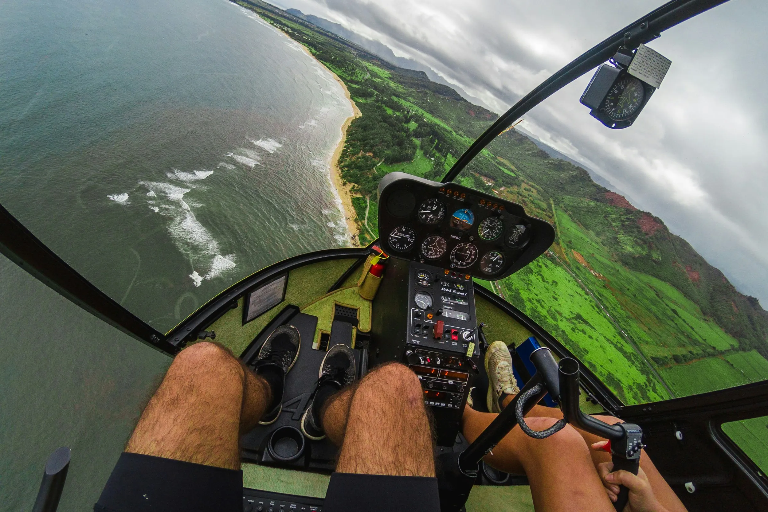 A helicopter cockpit view shows two people flying over coastline and green fields under a cloudy sky.