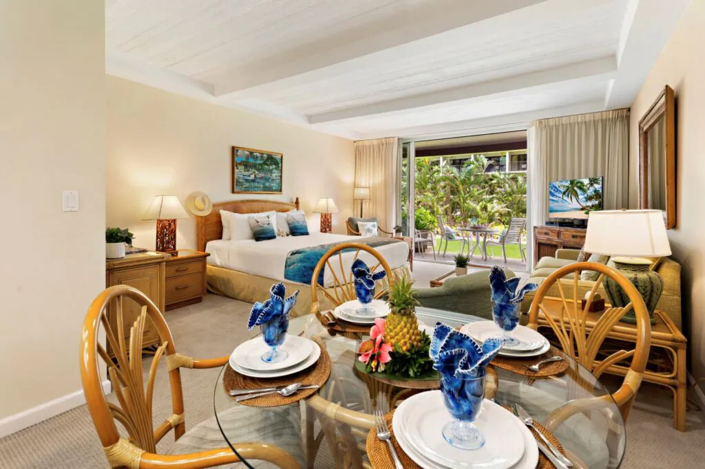 Bright tropical hotel room with a king bed, glass dining table set for four, TV, and a balcony overlooking lush greenery.