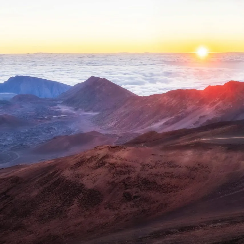 Sun rises above a sea of clouds and rugged volcanic mountains with reddish-brown slopes in a dramatic landscape.
