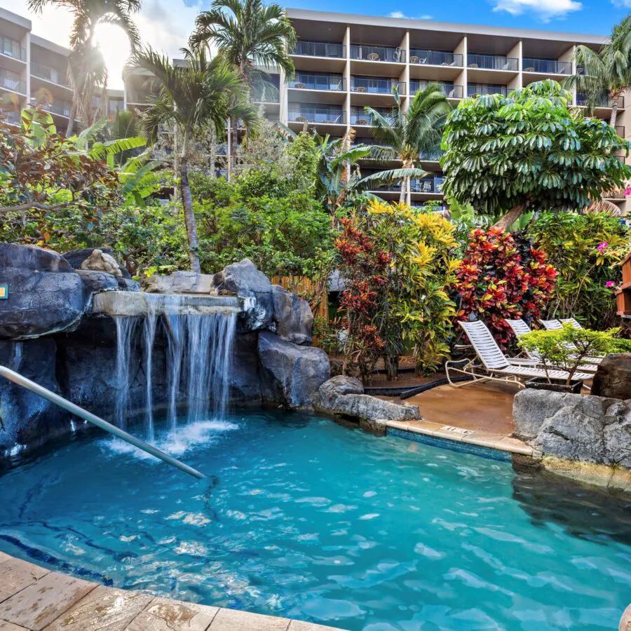 Small pool with a rock waterfall, surrounded by tropical plants and lounge chairs, in front of a multi-story resort building.
