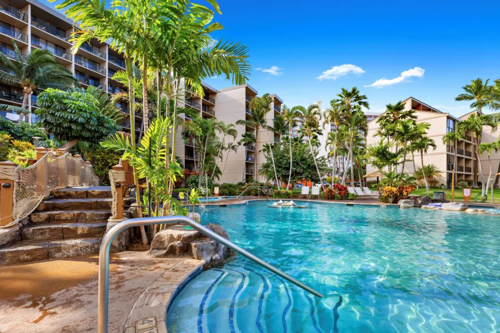 Resort pool surrounded by palm trees and tropical plants, with apartment buildings in the background under a bright blue sky.