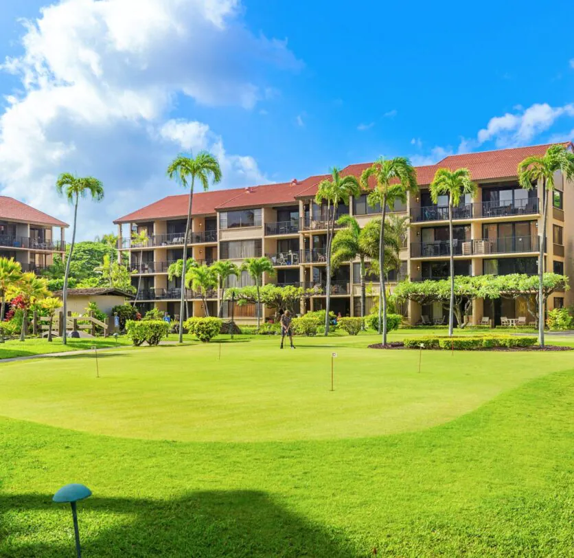 Sunny day at a resort with green lawns, palm trees, and multi-story buildings with balconies and red roofs under a bright blue sky.