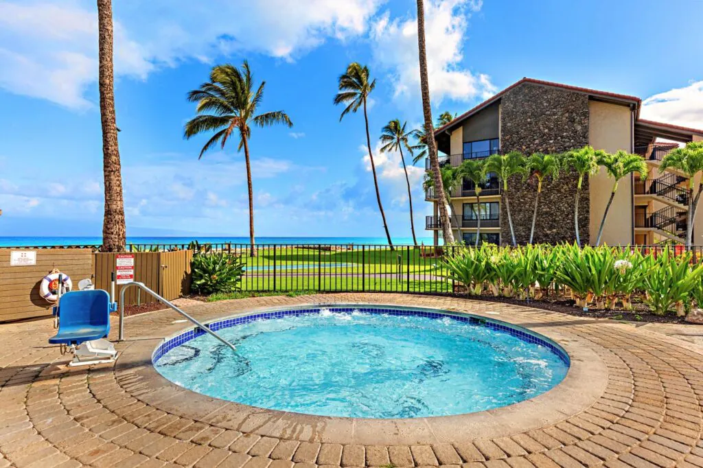 A round hot tub with blue water, palm trees, a fence, and a building nearby, overlooking a grassy area and the ocean under a blue sky.
