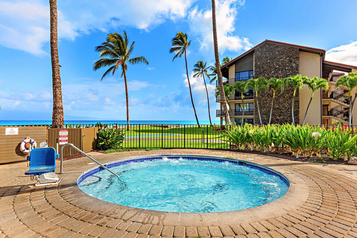 A round hot tub with blue water, palm trees, a fence, and a building nearby, overlooking a grassy area and the ocean under a blue sky.