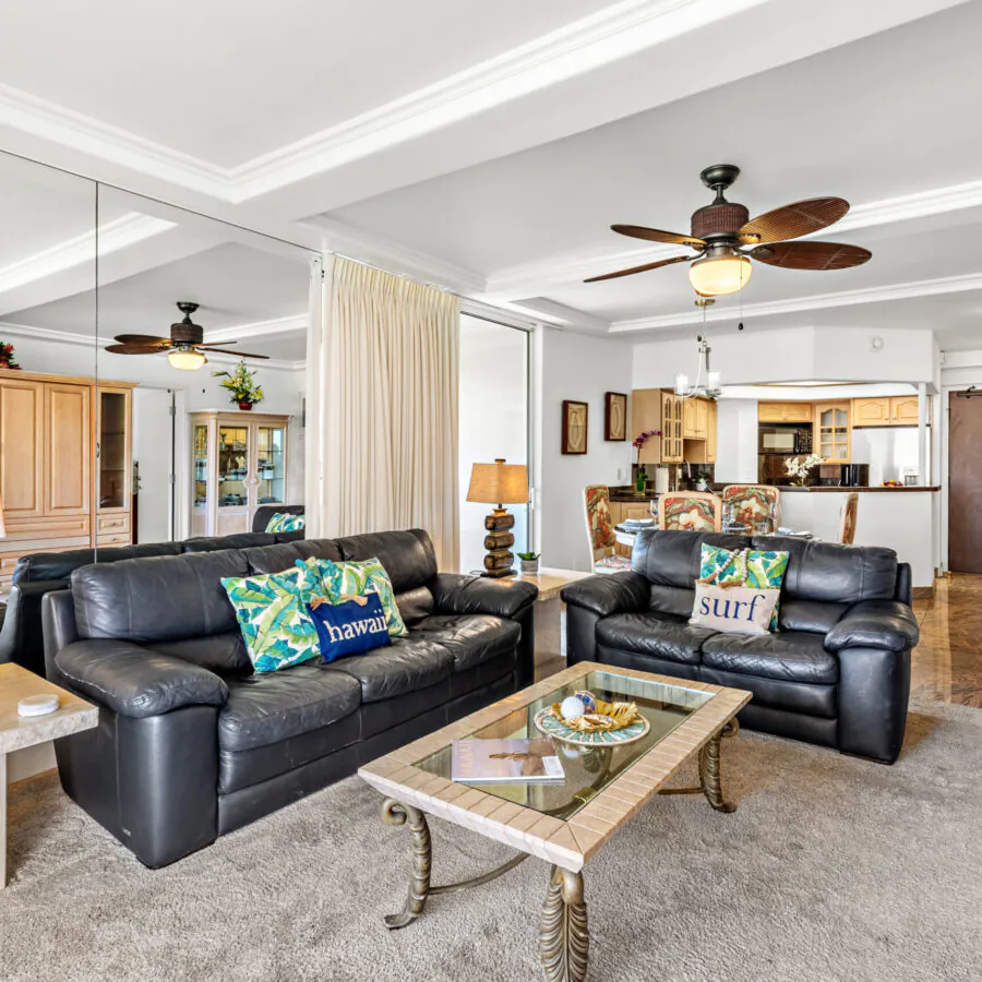Bright living room with black leather sofas, tropical-themed pillows, glass coffee table, and open view to kitchen with ceiling fans overhead.