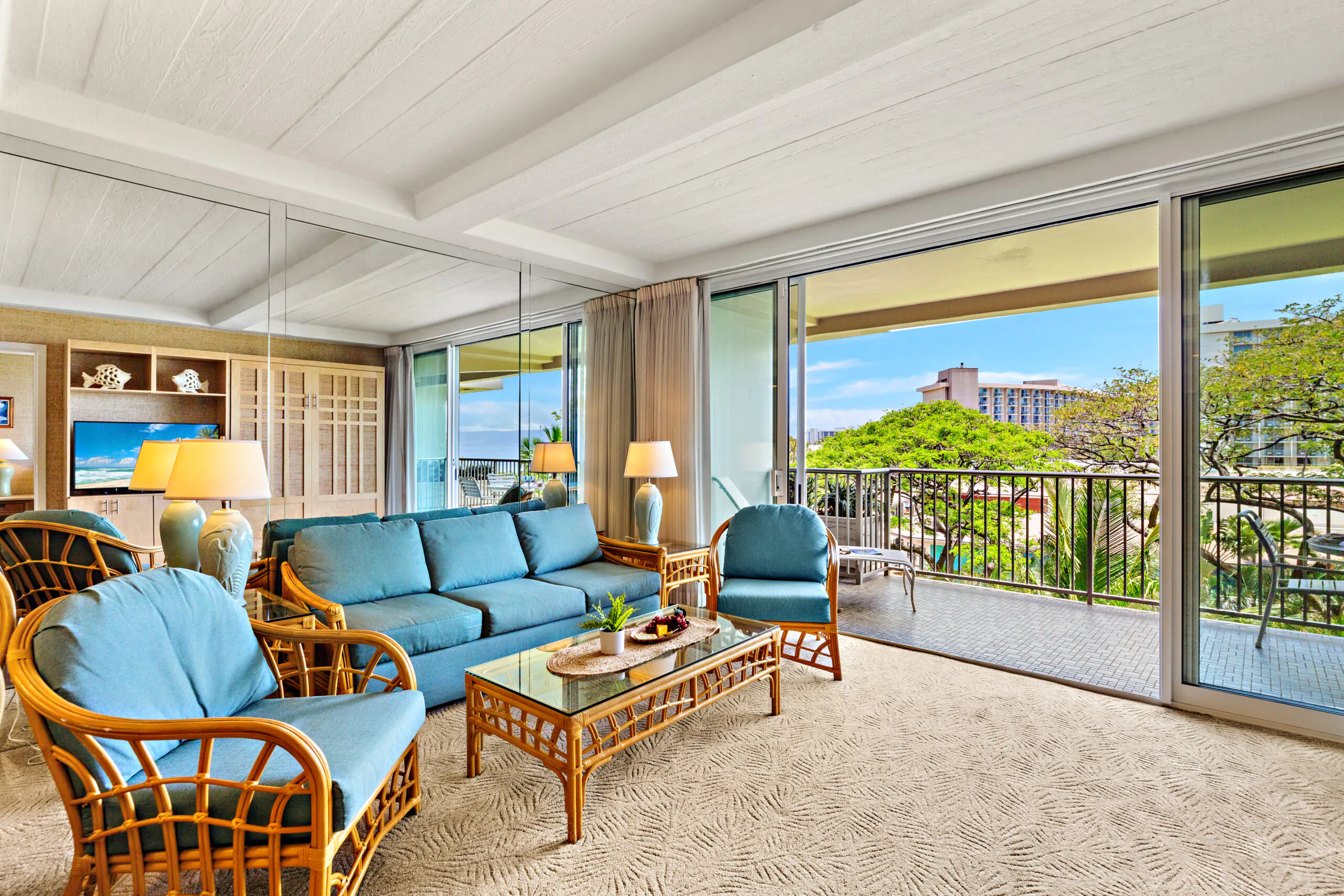 Bright living room with blue rattan furniture, glass coffee table, large windows, and a balcony overlooking trees and city buildings.