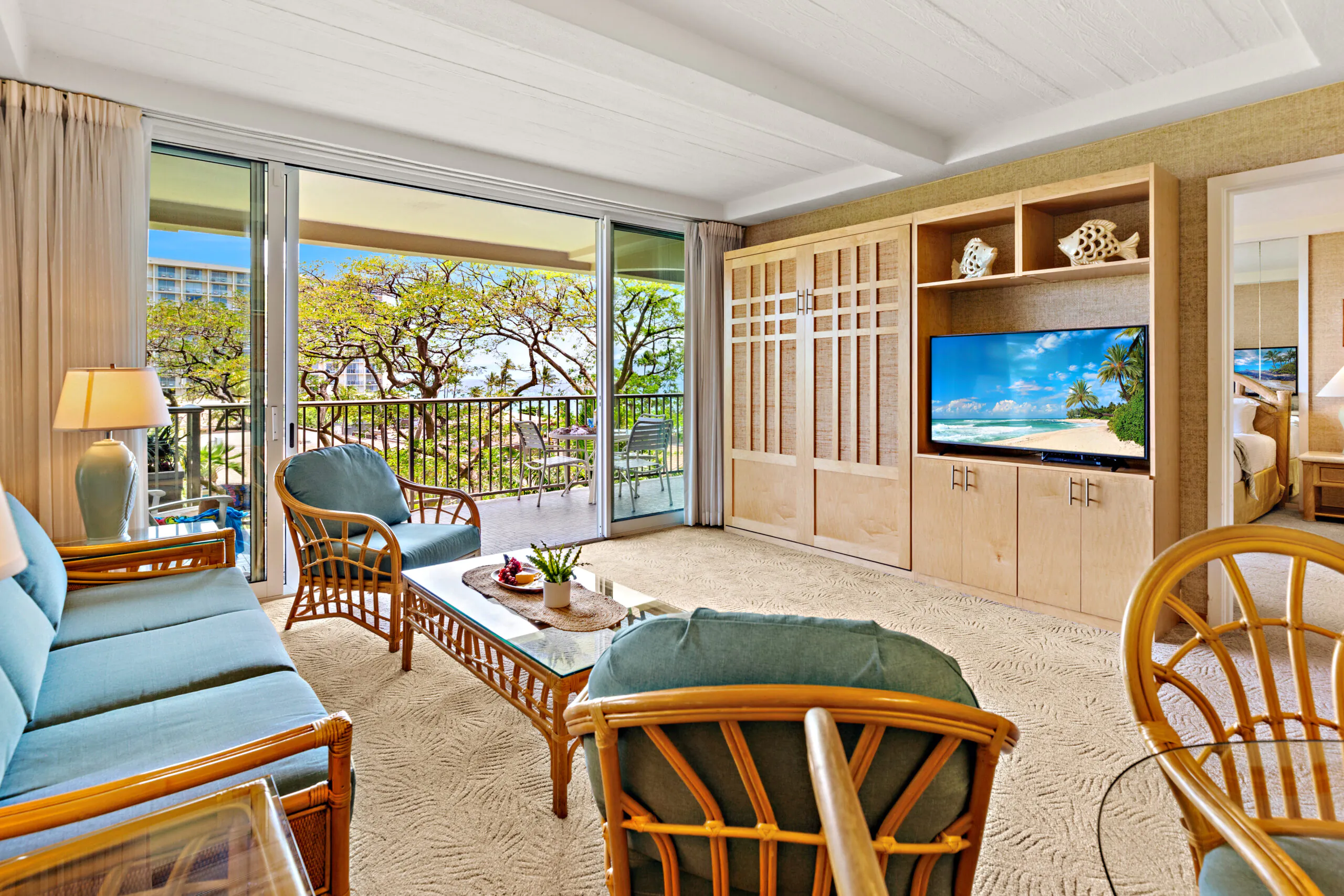 Bright living room with wicker furniture, a TV displaying a beach scene, large windows, and a balcony overlooking green trees.