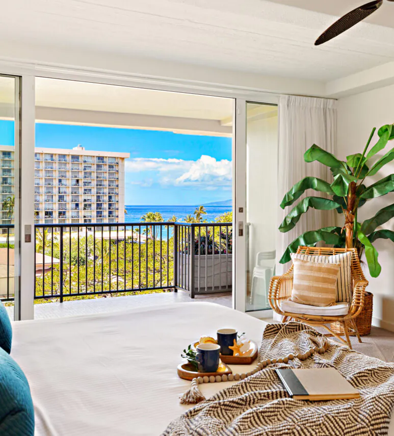 Bright bedroom with a king bed, tropical decor, balcony, ocean view, potted plant, and a tray with coffee and a book on the bed.