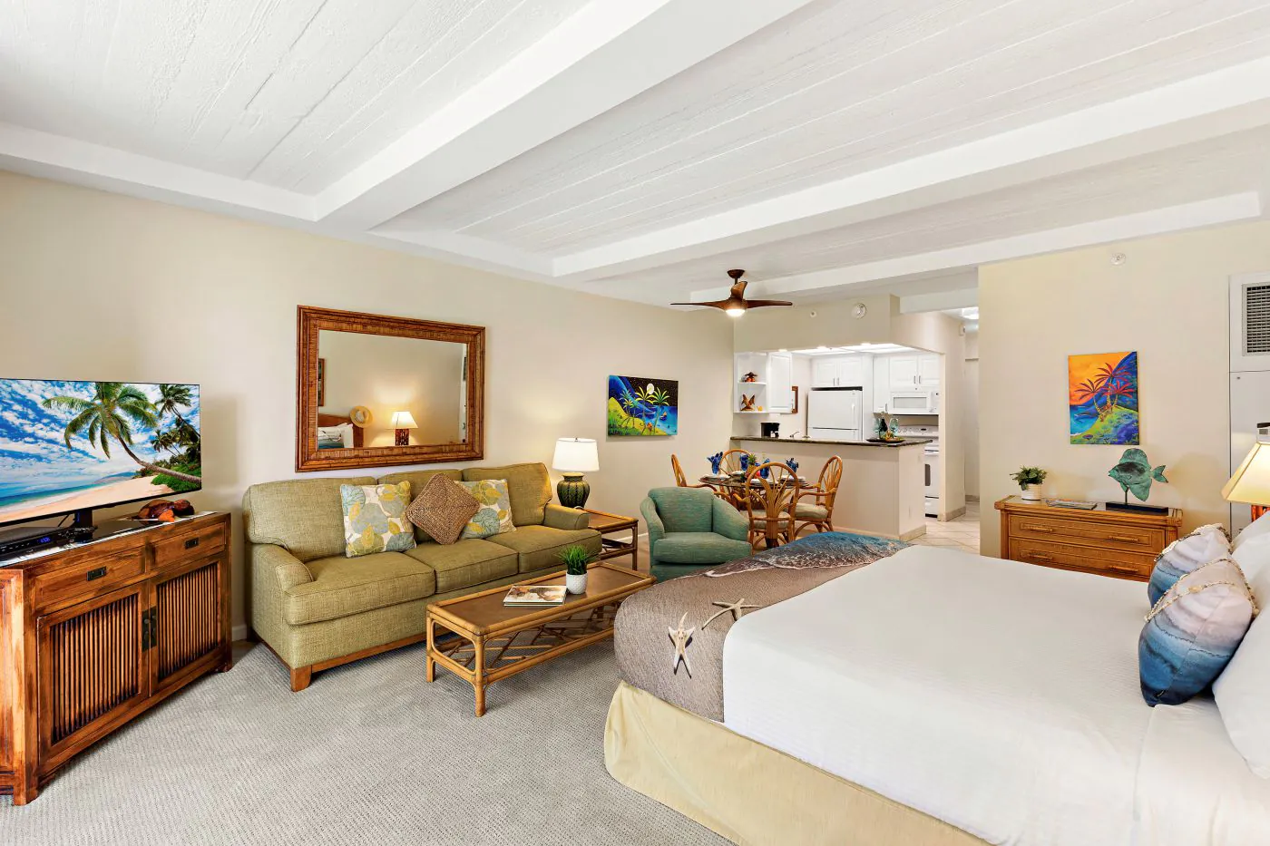 Bright, airy studio with a bed, sofa, TV, dining table, tropical decor, and a kitchenette in the background.
