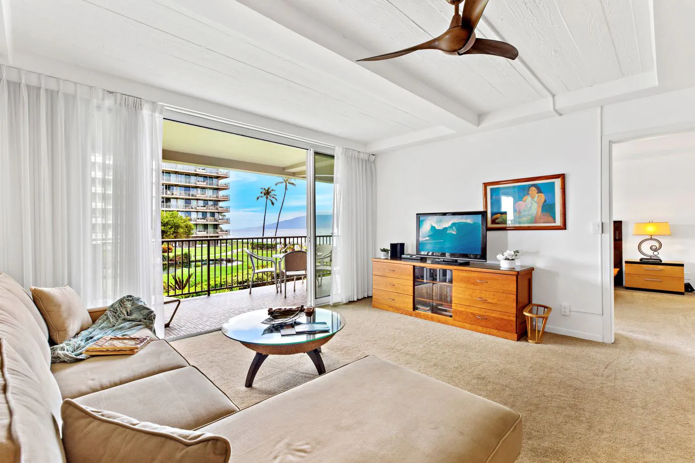 Bright living room with beige sofa, TV, wooden furniture, balcony view of palm trees, ocean, and adjacent bedroom.